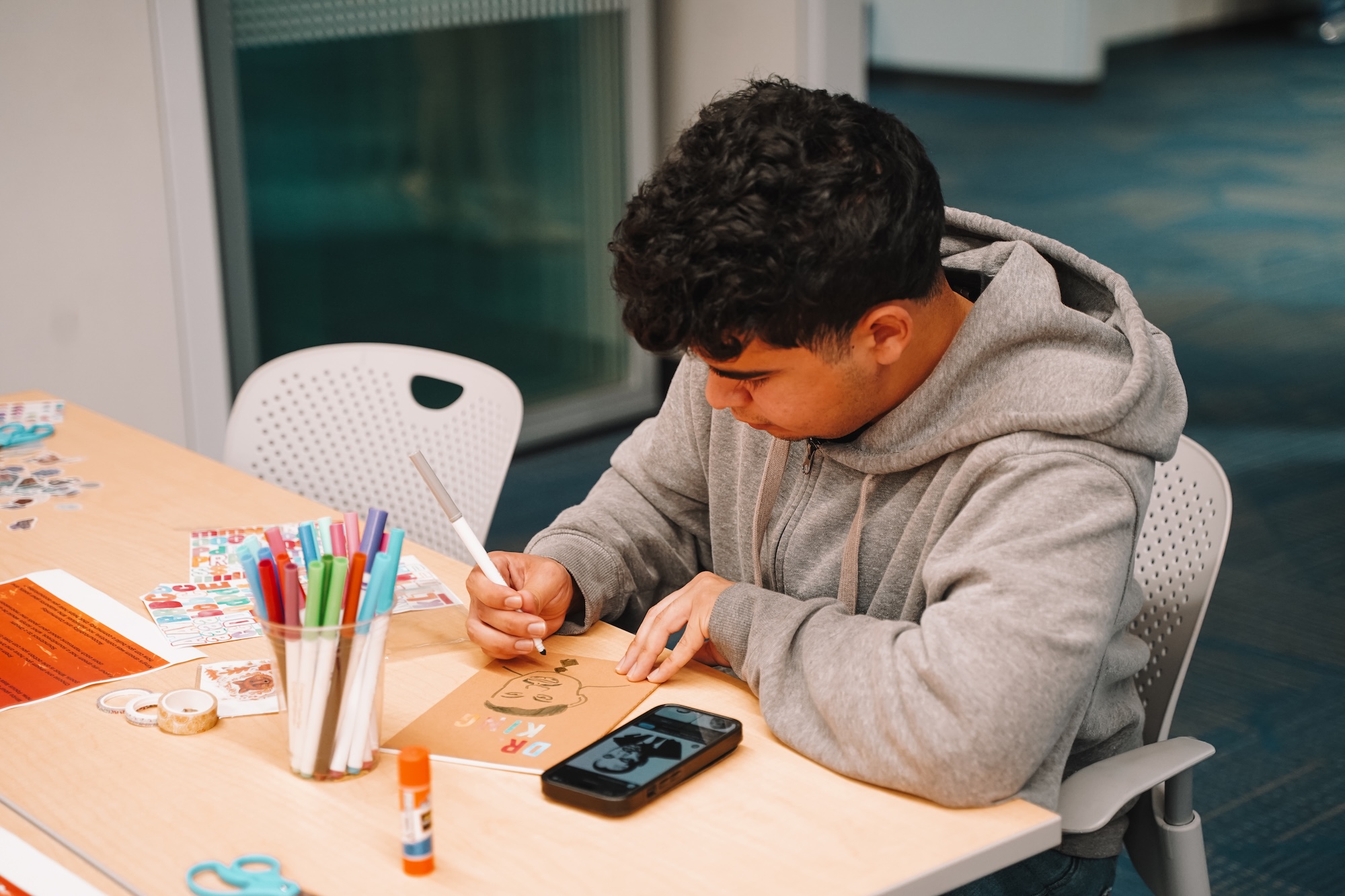 Student in a grey hoodie sitting at a table and drawing on the cover of his journal.